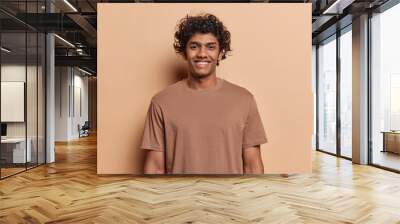 Portrait of happy Hindu man with curly hair smile toothily keeps arms down dressed in casual t shirt looks cheerful isolated over brown background being in good mood. Human positive emotions concept Wall mural