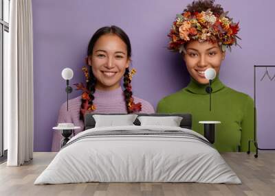 Portrait of happy mixed race women in warm clothes, hair decorated with autumn leaves, express good emotions, stand next to each other. Asian girl with two plaits, Afro lady wears seasonal wreath Wall mural