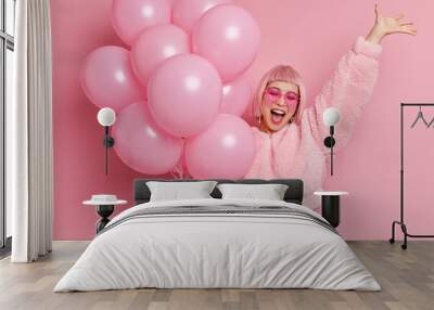 Portrait of joyful Asian woman with pink hair raises hand has fun exclaims happily finally passed all exams at university and has graduation party dressed in festive clothes holds bunch of balloons Wall mural