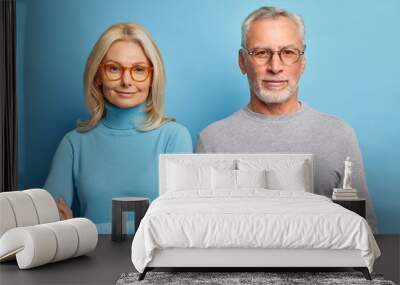 Portrait of mature woman and man stand next to each other in casual clothes against blue background look directly at camera with calm expressions. Senior couple pose indoor. People age concept Wall mural