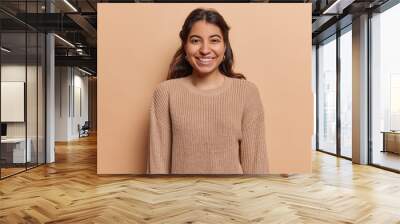 Portrait of pleasant looking Iranian girl with toothy smile has good mood dressed in casual knitted jumper looks directly at camera isolated over brown background. People and positive emotions concept Wall mural