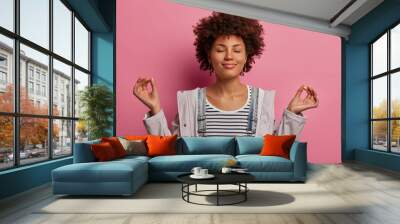 Portrait of relieved determined dark skinned woman meditates with closed eyes, practices breathing exercise or yoga to calm down, stands in lotus pose, poses over pink background, makes zen gesture Wall mural