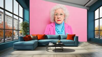 Portrait of serious mature grey haired woman dressed in elegant clothes with jewelry wears makeup remains beautiful despite her old age poses indoor against pink background. Fashionable grandmother Wall mural