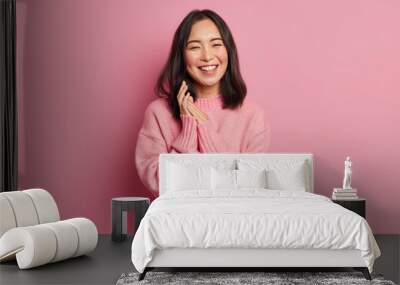 Portrait of sincere brunette Asian female model rubs palms and smiles broadly expresses happiness feels joyful has white even teeth wears oversized casual sweater isolated over pink background Wall mural