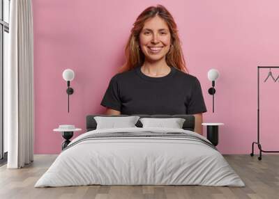 Portrait of smiling European woman being in good mood poses against pink background dressed in casual black t shirt. Beautiful female model expresses positive emotions stands indoor at studio Wall mural