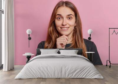 Portrait of surprised long haired woman keeps hand on chin bites lips looks with curious expression at camera wears casual black t shirt isolated over pink background. Human facial expressions Wall mural