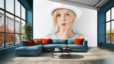 Portrait of surprised or frightened girl looking at the camera with a hand on her cheek. Close up shot of blonde Caucasian little girl with scared or shocked expression against white studio wall Wall mural