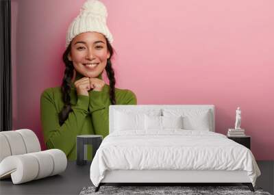 Portrait of tender lovely Asian woman has rouge cheeks, pleasant smile, keeps hands under chin, looks happily directly at camera, wears knitted hat, green jumper, being interested in nice idea Wall mural