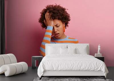 Portrait of unhealthy young curly woman has severe headache, keeps hand on forehead, feels unwell because of migraine, being stressed and worried, keeps eyes closed, isolated on pink background. Wall mural