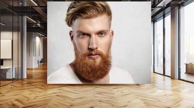 Portrait of young bearded dandy with hair brushed back in a trendy manner. Attractive Caucasian man in white T-shirt looking ahead, his charming green eyes sparkling. Indoors shot in white studio. Wall mural