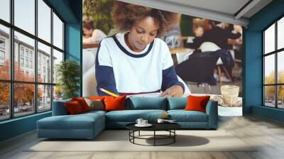 Portrait of young dark-skinned woman in casual clothes sitting at University canteen doing home assignment using modern gadget, studying something on tablet, drawing diagrams or graphs at work-book Wall mural