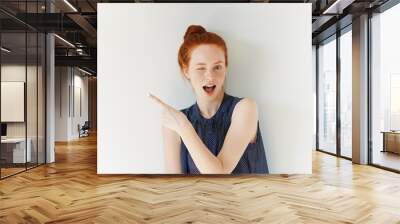 Portrait of young redhead female with perfect freckled skin showing something interesting at copy space wall for your information or advertising content, winking at the camera and pointing sideways Wall mural