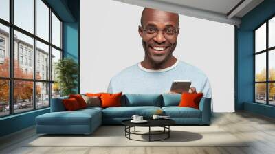 Positive African American male with broad smile, holds modern cell phone, types text message and feedback, surfes social networks, rejoices high speed interent connection, dressed in blue sweater Wall mural