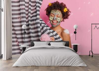 Positive Afro American woman sings song while showering applies cucumber facial mask to rejuvenate skin holds sponge enjoys body care procedures isolated on pink background soap bubbles flying around Wall mural