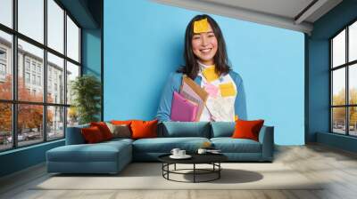 Positive Asian student holds folders stands glad with stickers on clothes and forehead prepares project work in economics happy to finish doing important task wears jumper isolated on blue wall Wall mural