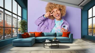 Positive carefree young woman with curly hair makes peace gesture over eye smiles happily holds mobile phone sends text messages dressed in cropped top and jacket isolated over purple background Wall mural