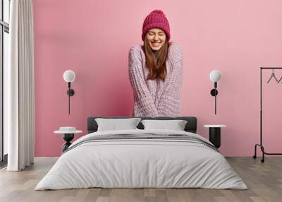 Positive Caucasian young female model keeps hands together, wears long sleeved knitted sweater, pink hat, closes eyes from pleasure, models over rosy background. People and emotions concept. Wall mural