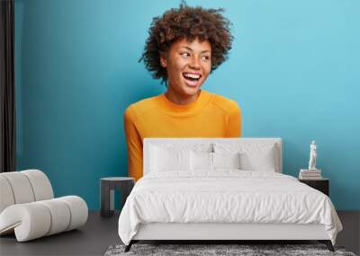 Positive cheerful young African American woman laughs positively and looks aside concentrated happily dressed in casual orange jumper isolated on blue studio background. People emotions concept Wall mural