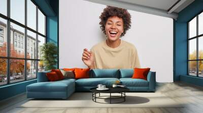 Positive curly haired beautiful woman laughs happily has carefree expression keeps mouth opened wears casual beige t shirt isolated over white studio background. Sincere human emotions concept Wall mural