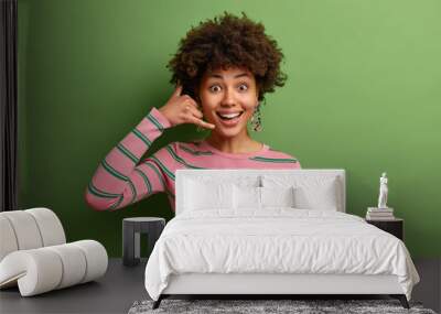 Positive dark skinned curly woman makes call me gesture keeps hands shaped like phone wears striped jumper poses against green background. Cheerful ethnic lady asks for your telephone number Wall mural