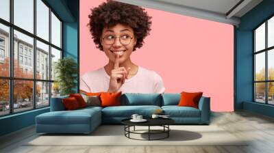 Positive dark skinned female with curly bushy hair, has toothy smile, makes hush gesture, wears eyeglasses and casual clothes, poses against pink studio wall with copy space for your advertisement Wall mural