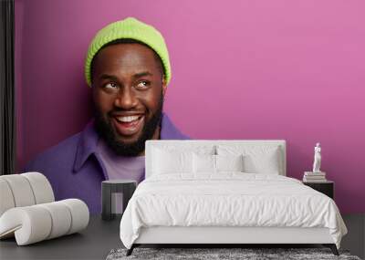 Positive dark skinned man remembers good moment, laughs and looks somewhere pensively, has happy reaction, dressed in bright clothes, stands indoor, blank space for your advertising content. Wall mural