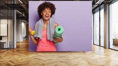 Positive dark skinned woman holds yoga mat and dumbbell, wears sportclothes, has towel on neck, looks aside with happy expression, exercises in gym, isolated over purple wall. Fitness and training Wall mural