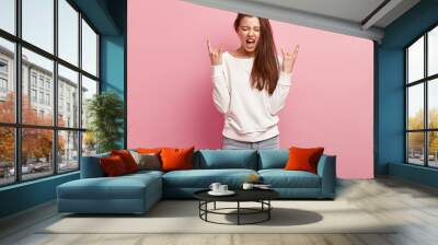 Positive emotional woman makes rock n roll gesture, exclaims loudly while listens loud music, dressed in casual jumper and jeans, isolated over pink background. I am fun of rock. Horizontal shot Wall mural