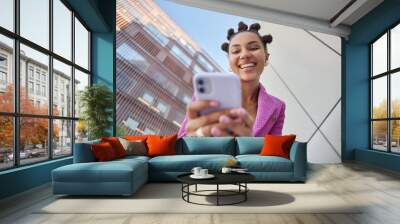 Positive fashionable woman uses mobile phone for chatting sends text messages surfs social networks dressed in pink jacket poses against urban background strolls in city has trendy hairstyle Wall mural