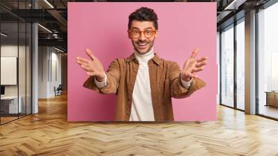 Positive friendly European man raises hands as meets someone pulls arms in welcome gesture wants to embrace friend smiles gladfully wears stylish shirt and spectacles isolated over pink background Wall mural