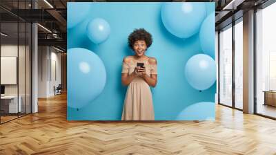 Positive glad African American woman holds mobile phone in hands and happy to chat with friends in social networks, wears cocktail dress, poses against blue background in decorated photo zone Wall mural