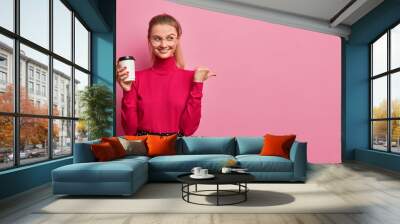 Positive good looking girl with combed tail smiles gladfully and points away on empty space drinks takeaway coffee dressed in casual wear isolated over pink background gives way to cafe or coffee shop Wall mural