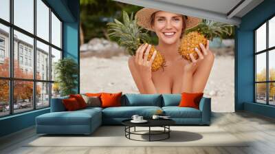 Positive half nude female with cheerful smile wears only straw hat, holds two big pineapples in hands, poses on sandy beach. Adorable woman vegeterian eats low calorie food. Summer time concept Wall mural