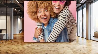 Positive happy women of different race have fun together look gladfully and surprised dressed in casual clothes isolated over pink background. Two female friends embrace foolish around indoors Wall mural