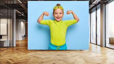 Positive little girl spends leisure time in water park, raises arms and shows muscles, ready for swim and dive, poses over blue background, wears goggles and swimsuit. Small kid has healthy lifestyle Wall mural