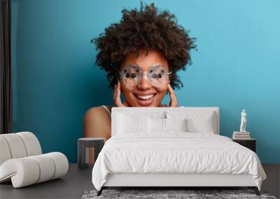 Positive relaxed curly haired woman wears rubber waterproof goggles on eyes, ready for taking shower, touches cheeks gently, smiles broadly, isolated on blue background. People, lifestyle, hygiene Wall mural