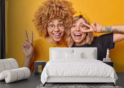 Positive two female friends show peace gesture smile gladfully laugh happily dressed in casual t shirts isolated over yellow background. Playful girls go crazy have good mood make victory sign Wall mural