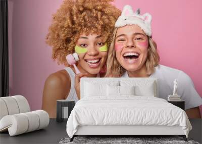 Positive two women apply beauty facial mask and pads under eyes use face massager smile broadly dressed in casual t shirts being in good mood isolated over pink background. Wellness concept. Wall mural