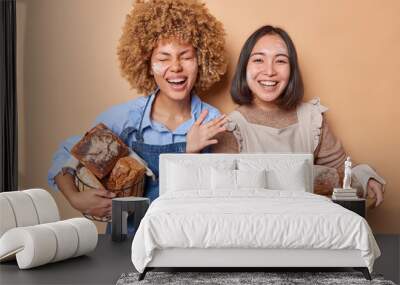 Positive two women stand happy with fresh delicious bread smeared with flour wear aprons satisfied after finishing baking isolated over brown background. Female bakers work at bakery factory Wall mural