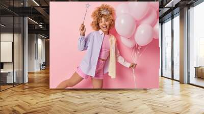 Positive woman has fun before holiday holds bunch of inflated balloons and magic wand stands on one leg enjoys party time poses against pink background has festive mood. Special occasion concept Wall mural