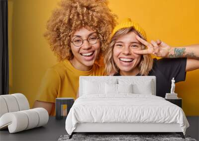 Positive women have joyful expressions smile broadly make peace gesture laugh at something funny foolish around dressed casually isolated over vivid yellow background. Happy emotions concept Wall mural