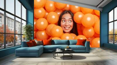 Positive young Asian woman smiles broadly and looks away surrounded by many orange air balloons enjoys birthday party event has long dark hair celebrates something being in good festive mood Wall mural