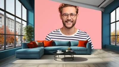 Positive young Caucasian male with pleasant friendly smile, shows white teeth, rejoices new stage in life, wears casual striped sweater and round spectacles, stands alone against pink background. Wall mural