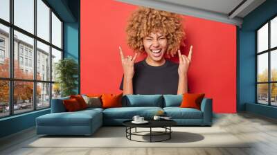 Positive young curly woman shows horn gesture enjoys rock music wears black casual t shirt isolated over vivid red background. Overjoyed curly haired female model demonstrates heavy metal sign Wall mural