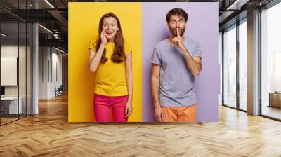 Positive young female whispers secret with glad expression, serious guy keeps fore finger on lips, gossips and spread private information, wear casual t shirts, stands against colorful background Wall mural