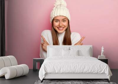 Positive young girl with straight hair, crosses arms and points in both sides, wears fashionable white hat and jumper, chooses between two items, smiles pleasantly, isolated over pink background. Wall mural