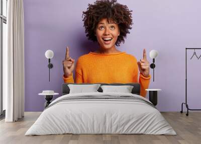 Positive young good looking woman with Afro hairstyle, indicates with both index fingers, wears casual orange sweater, feels pleased, isolated over purple background. People and promotion concept Wall mural