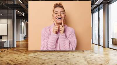 Positive young woman with combed hair points at her broad toothy smile feels very happy has white perfect teeth wears knitted sweater isolated over beige background. People and emotions concept Wall mural