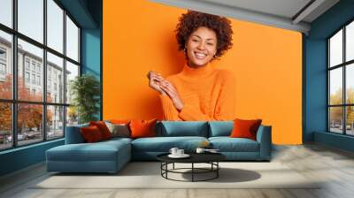 Pretty curly haired young woman smiles gently keeps hands together focused directly at camera has pleasant talk with best friend dressed in casual sweater isolated over vivid orange background Wall mural