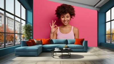 Pretty dark skinned female model shows okay gesture, reassures everything goes excellent, demonstrates no problem sign, gives positive feedback or likes idea, wears casual tank top, isolated on pink Wall mural